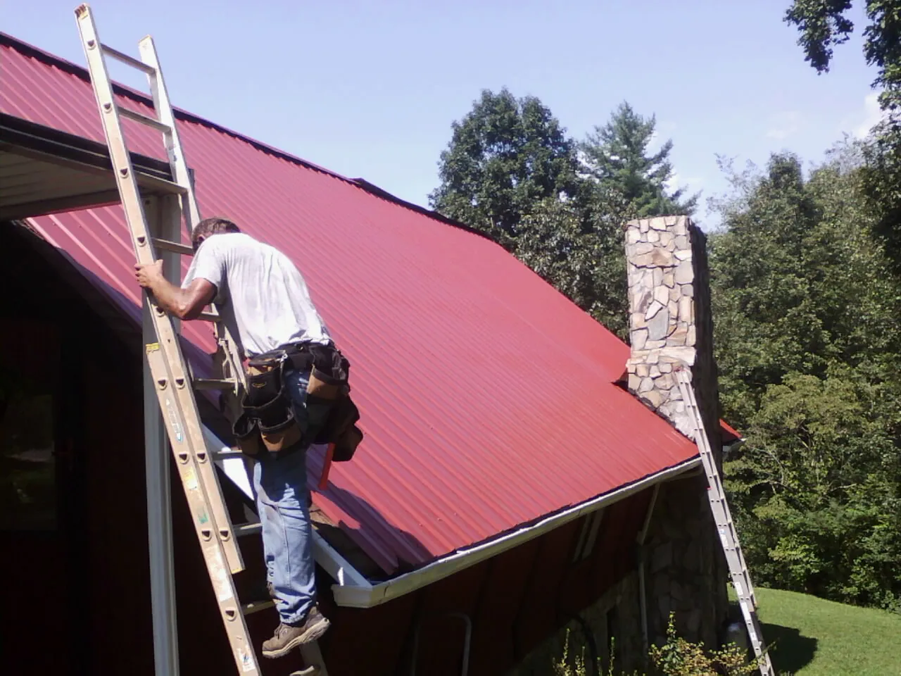 Roofer on a ladder beside a red metal roof and stone chimney — Soffit & Fascia Repair by Roofing Brothers in Conesville